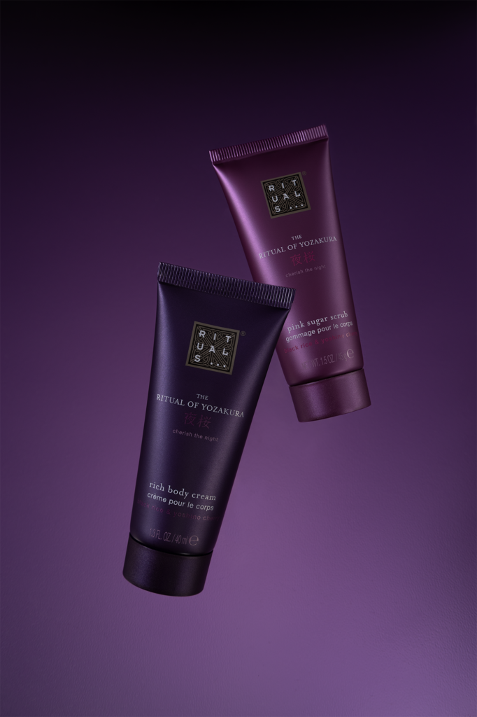 Professional product photography of Rituals The Ritual of Yozakura cosmetics. Two purple tubes of body cream and scrub levitating in the air against a dark purple gradient background with elegant studio lighting.